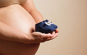 Blaue Babyschühchen werden vor einem runden Schwangerschaftsbauch gehalten.